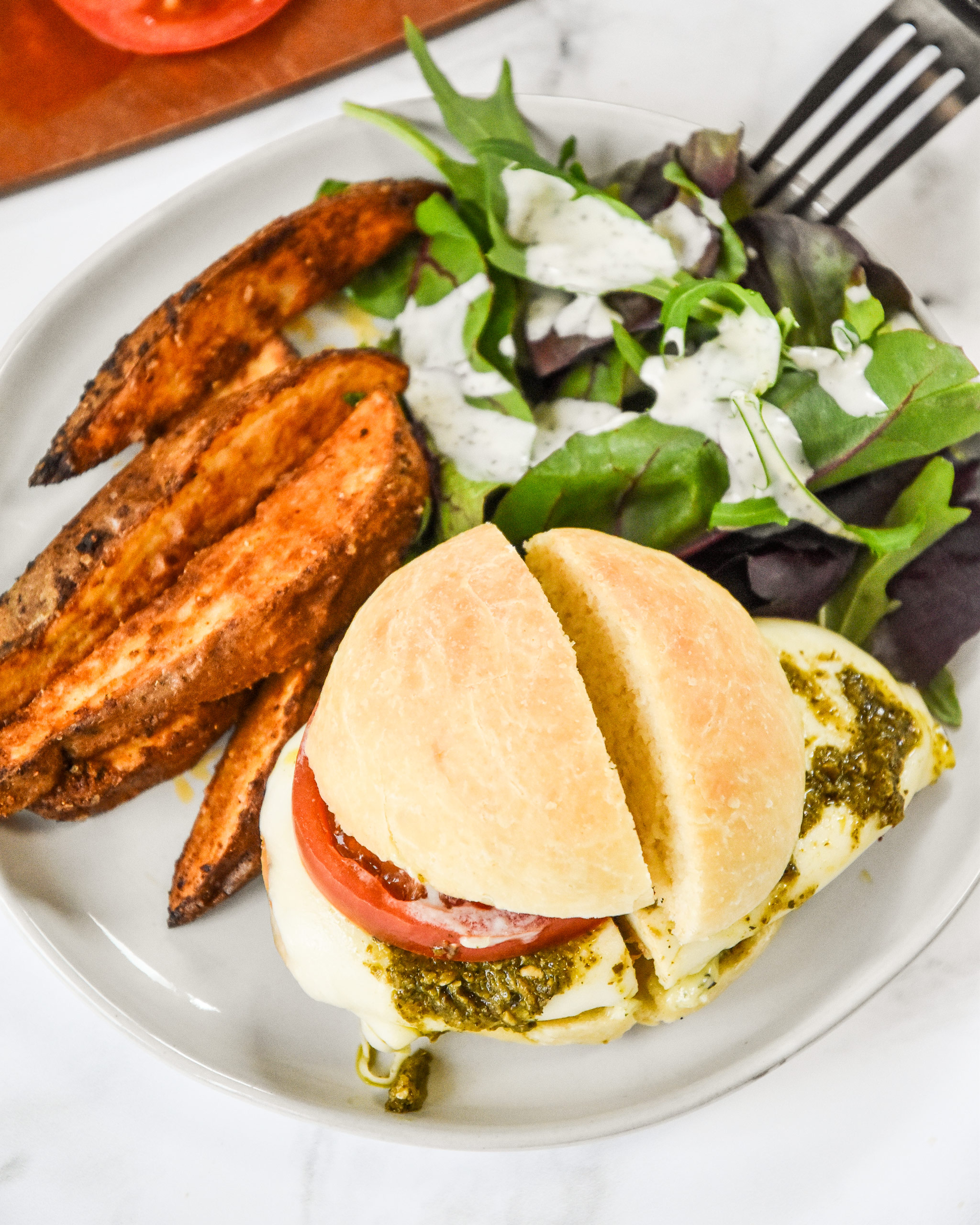 pesto chicken mozzarella sandwich on a plate with potato wedges and a salad.