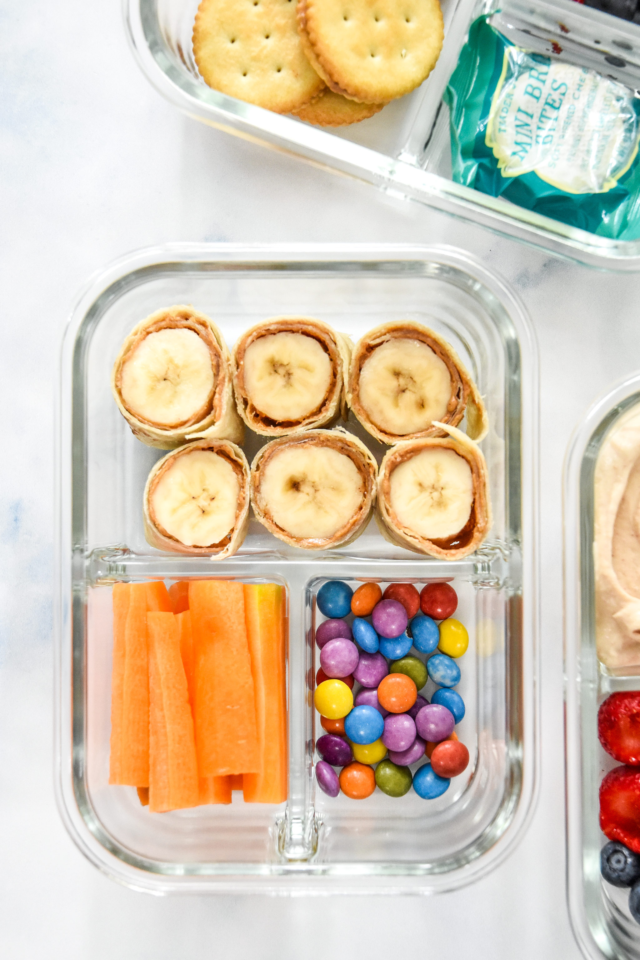 banana roll ups with carrot sticks and chocolate candies in a glass meal prep container.