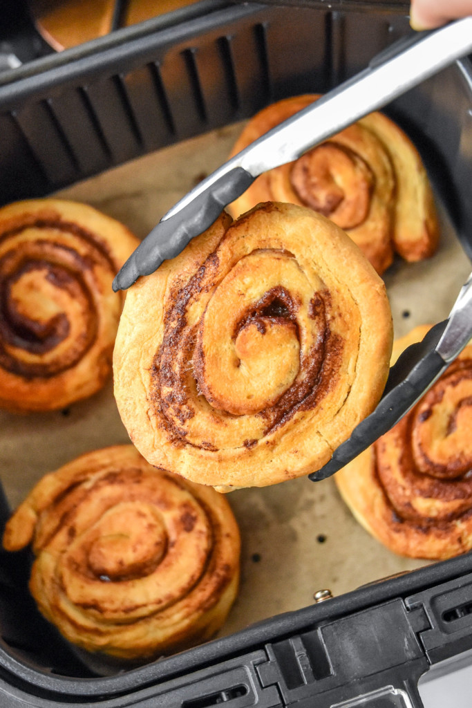 holding an air fryer canned cinnamon roll in tongs after cooking.