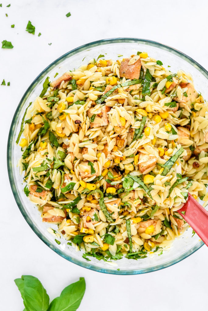 lemony chicken and corn orzo salad mixed up in a glass bowl with rubber spatula.