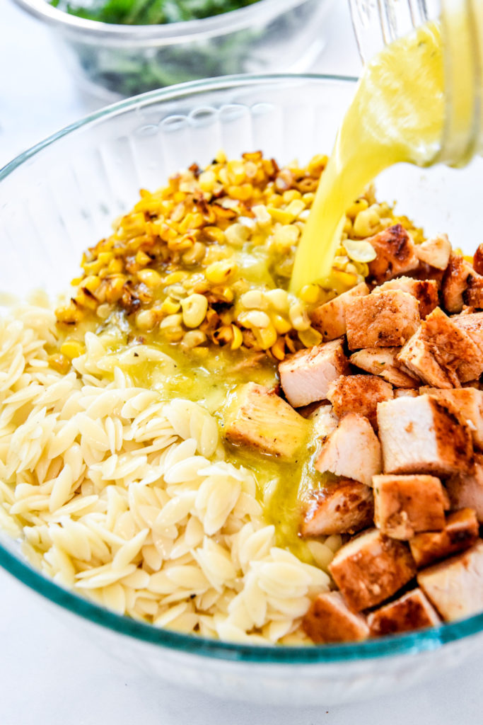 pouring lemon dressing into a bowl of orzo, chicken and corn.