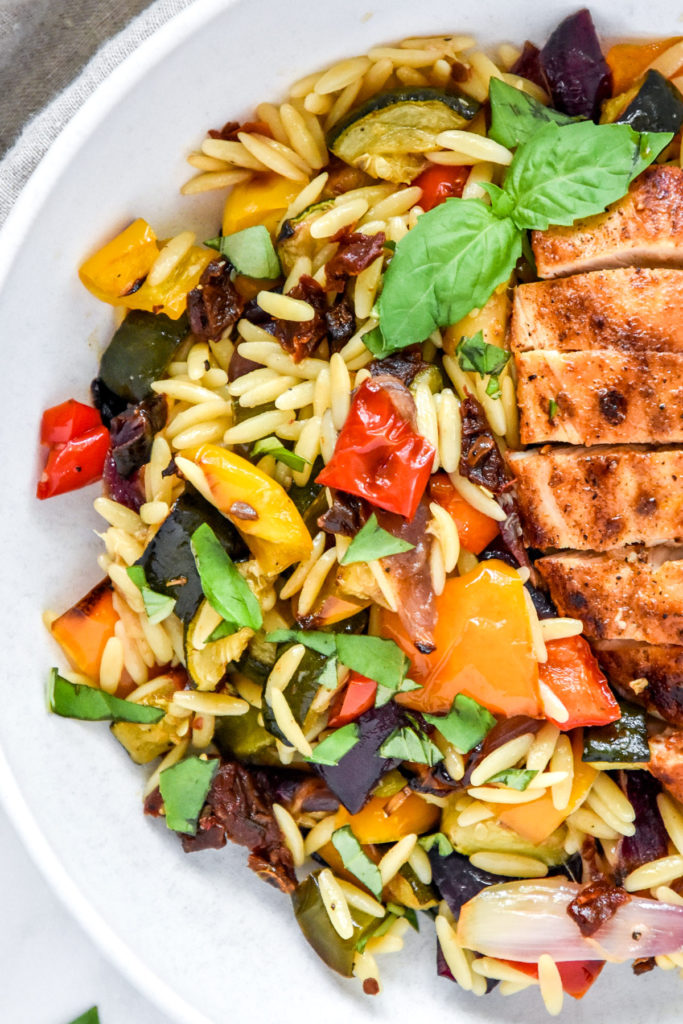 roasted vegetable orzo salad with sliced chicken breast in a white bowl.