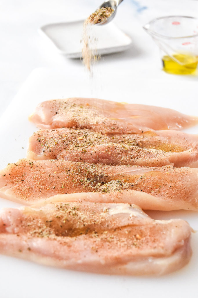 sprinkling seasoning on top of thin sliced chicken breasts.