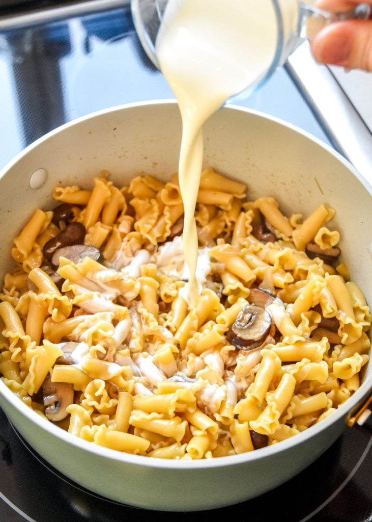 pouring heavy cream into a pot of cooked mushroom pasta.