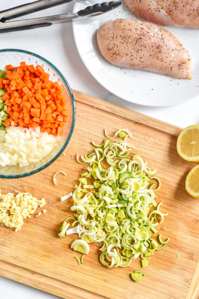 sliced leeks on a cutting board with other vegetables and seasoned chicken breast on a plate. 