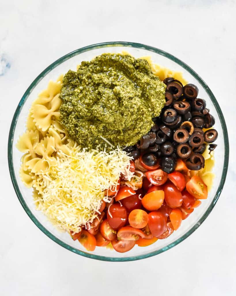 pesto, olives noodles tomatoes and parmesan cheese before stirring the pasta salad.