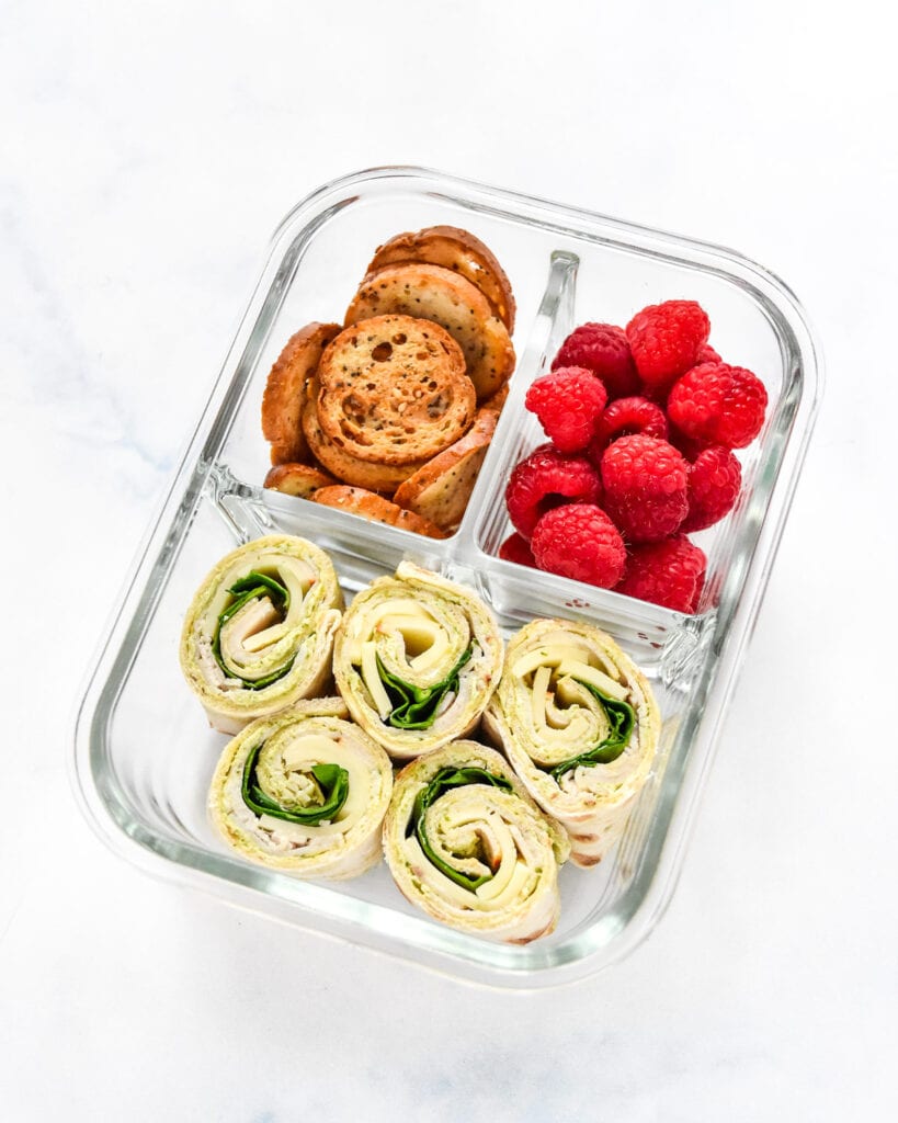 turkey pesto pinwheels in a glass meal prep container with raspberries and bagel chips.