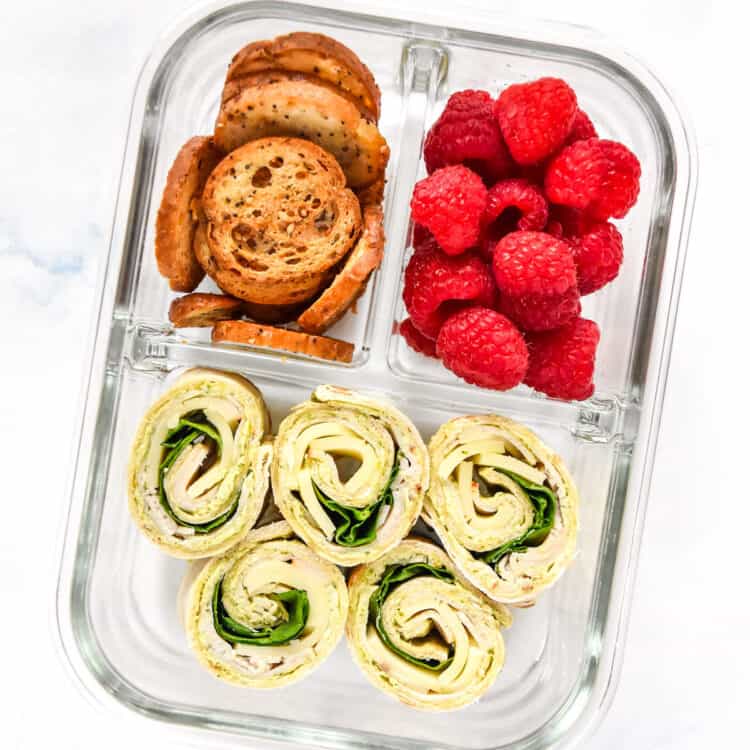 turkey pesto pinwheels in a glass meal prep container with raspberries and bagel chips.