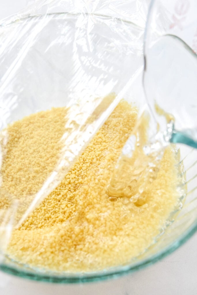 pouring hot water into a bowl of dry couscous with plastic wrap on top.
