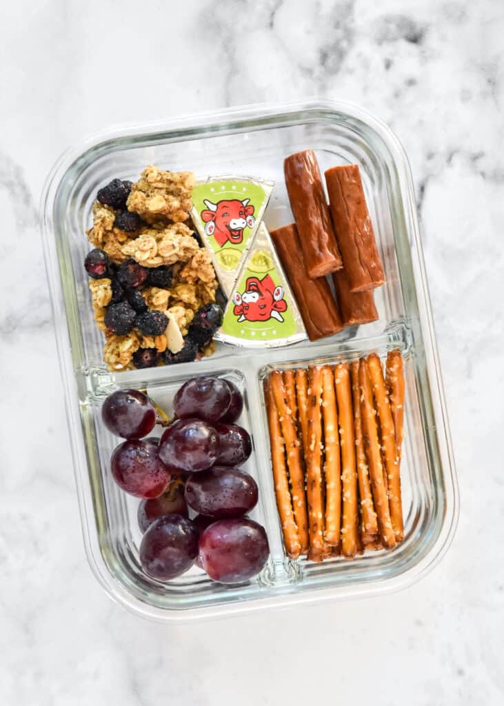 DIY adult lunchable with grapes, pretzels, meat stick, granola in a glass meal prep container.